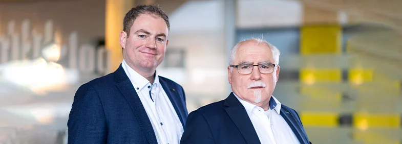 Florian Allertseder und Hans-Georg Allertseder im dunklen Sakko vor dem gelb-hölzernen Eingangsbereich der Kanzlei
